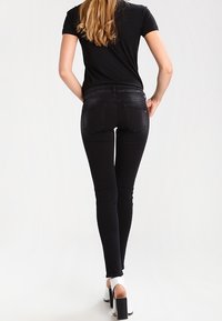 Jeans skinny negros de denim, con tiro alto, bolsillos traseros y una textura suave, combinados con sandalias blancas de tacón cuadrado.