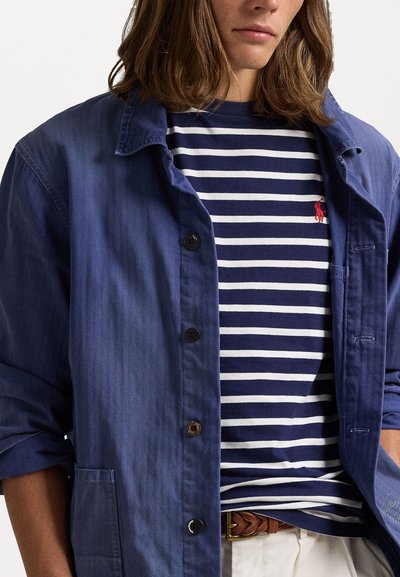 Námornícky modrá košeľa s gombíkmi z texturovanej látky, prekrývajúca pruhované námornícky modré a biele tričko. Je viditeľné malé červené logo.