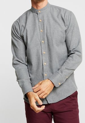 Man draagt een grijs overhemd met lange mouwen en mandarijnkraag met lichtbruine knopen en bordeauxrode broek, hij stelt zijn linkermanchet bij.