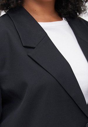 Nahaufnahme einer Person, die ein schwarzes Sakko über einem weißen, gerippten Hemd trägt, wobei der Kragen und der obere Brustbereich sichtbar sind.