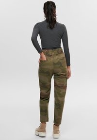 Frau mit dunklem Haar im Pferdeschwanz, die ein graues langärmliges Oberteil, grüne Camouflage-Hosen und beige Stiefel trägt, steht mit dem Rücken zur Kamera und hat eine Hand in der hinteren Tasche.