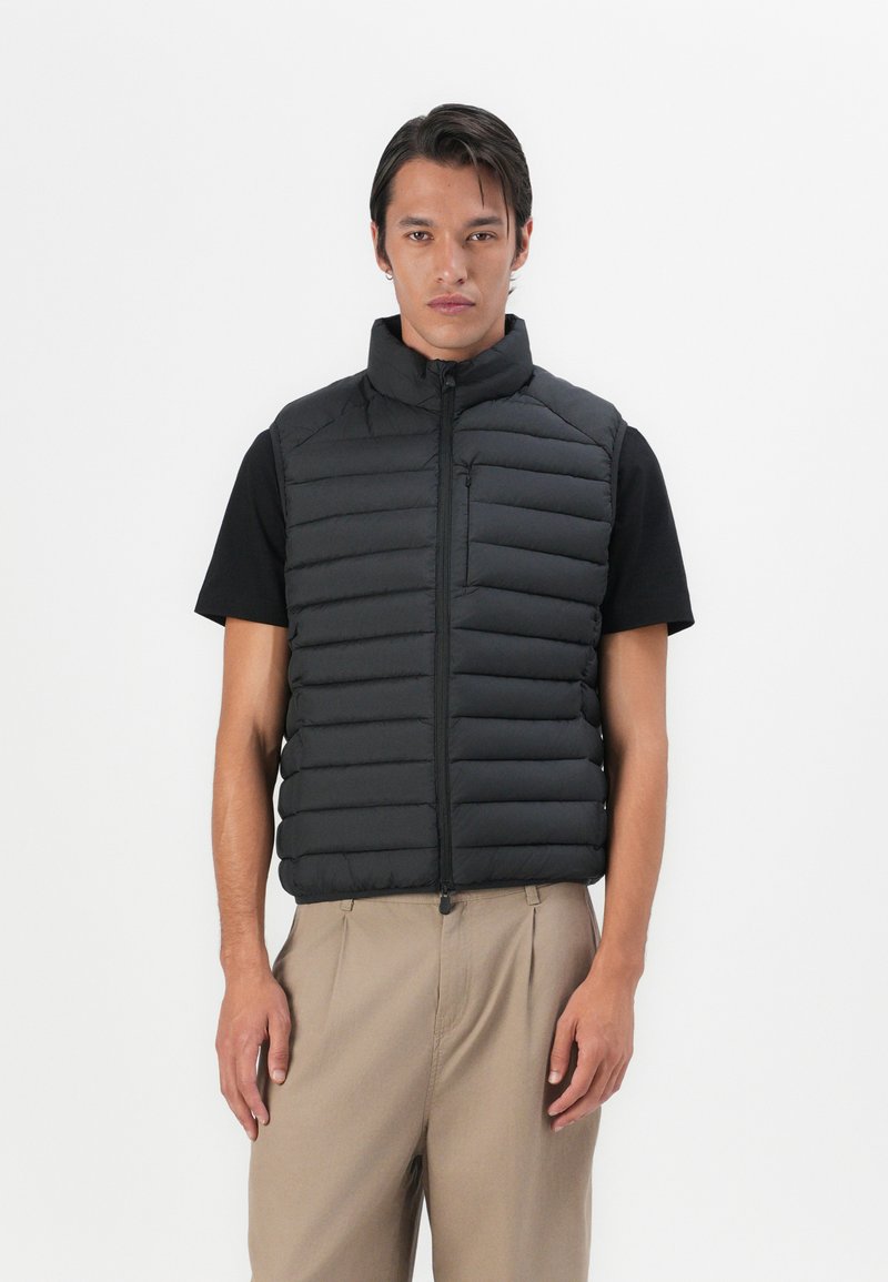 Sort puffer vest med horisontale syninger, høj krave, lynlåslukning og forlommer. Båret over en sort kortærmet skjorte og beige bukser.