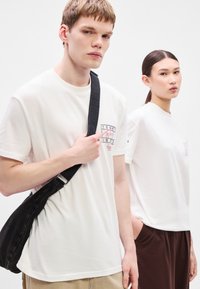 Kaksi henkilöä, jotka pukeutuvat valkoisiin puuvillaisiin t-paitoihin. Miehellä on t-paidassaan graafinen kuvio, kun taas naisella t-paita on yksinkertainen. Molemmat ovat rentoja malleja.