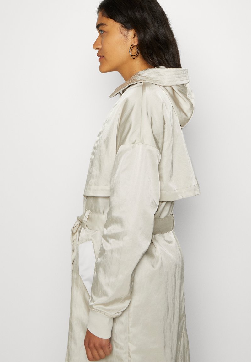 Trench coat de satén gris claro con capucha, mangas recortadas, cintura ceñida con cinturón y un bolsillo lateral con un acento blanco. Textura suave.