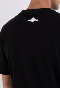 Camiseta de algodón negra con un logo blanco de corona y escudo en la parte superior de la espalda. Diseño simple con un cuello redondo clásico y textura suave.