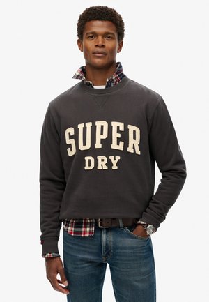 Sudadera gris con "SUPER DRY" en letras crema, superpuesta sobre una camisa de cuadros con tonos rojos, verdes y azules. Combinada con jeans azules.