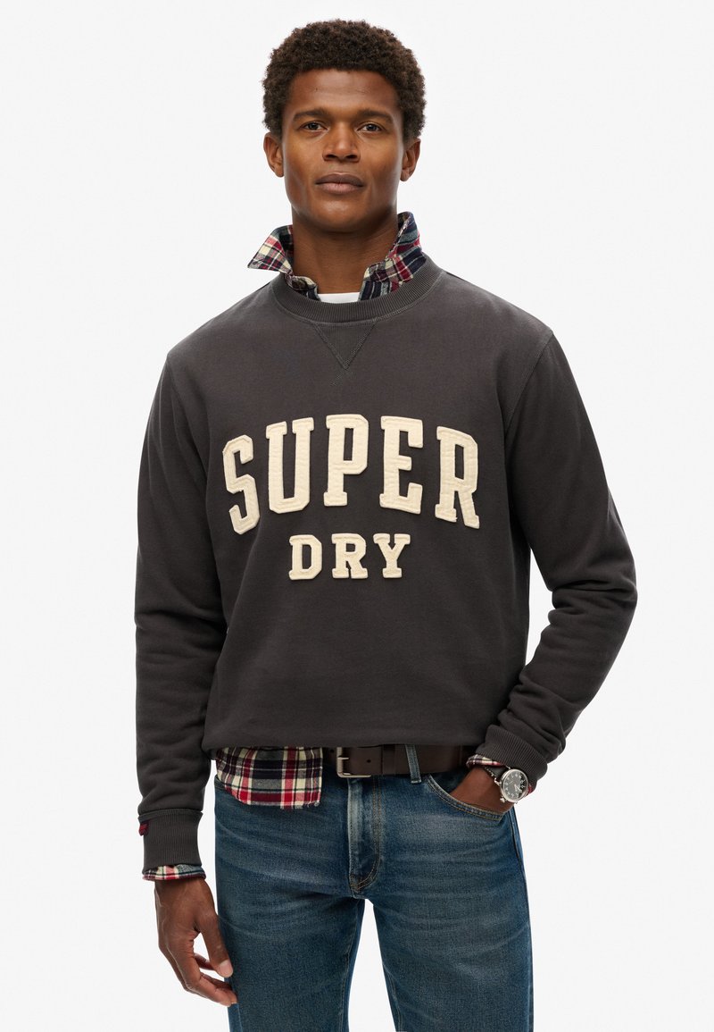Sudadera gris con "SUPER DRY" en letras crema, superpuesta sobre una camisa de cuadros con tonos rojos, verdes y azules. Combinada con jeans azules.