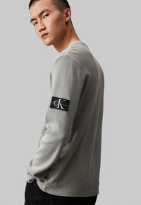 Camisa de manga larga gris con un tejido de punto texturizado, que presenta un parche negro con el logo de Calvin Klein Jeans en el brazo izquierdo.