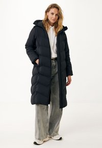 Černá péřová bunda s kapucí, s prošívaným designem, dlouhá a s bočními kapsami. Nosená přes bílé tričko a široké džíny.