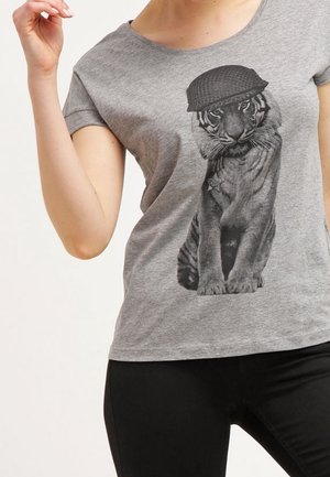 Kobieta ubrana w szary t-shirt z czarno-białym nadrukiem młodego tygrysa w hełmie wojskowym i z medalem na piersi.