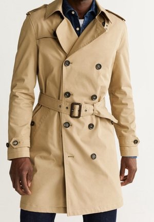 Homme portant un trench-coat beige croisé avec ceinture, poignets boutonnés et boutons foncés sur une chemise bleue et un sous-vêtement blanc.