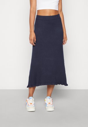 Kvinde iført en marineblå ribbet midi-nederdel med flæsekanter, hvid crop top og flerfarvede hvide sneakers, stående på en grå gulv.