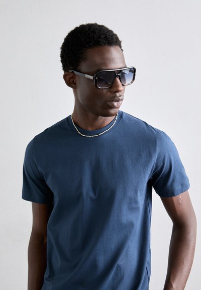 T-shirt en coton bleu marine avec un col rond et des manches courtes. Modèle portant de grandes lunettes de soleil noires angulaires et un collier en chaîne dorée fine.
