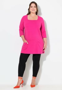 Fuchsia Tunika-Kleid mit quadratischem Ausschnitt, drei Viertelärmel und zwei vorderen Taschen, kombiniert mit schwarzen Leggings und orangenen Absatzschuhen.
