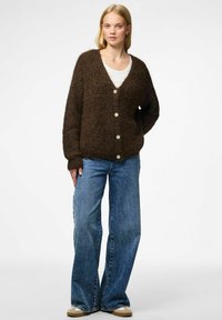 Blond naine, kes kannab pruuni karvase nupuga kardiganit valge pluusi, laia säärega siniste teksade ja beežide tossudega, valge tausta ees.