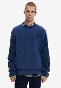 Blue oversized sweatshirt with textured cable knit sleeves, casual fit, and a ribbed cuff. Layered over a checkered shirt.
