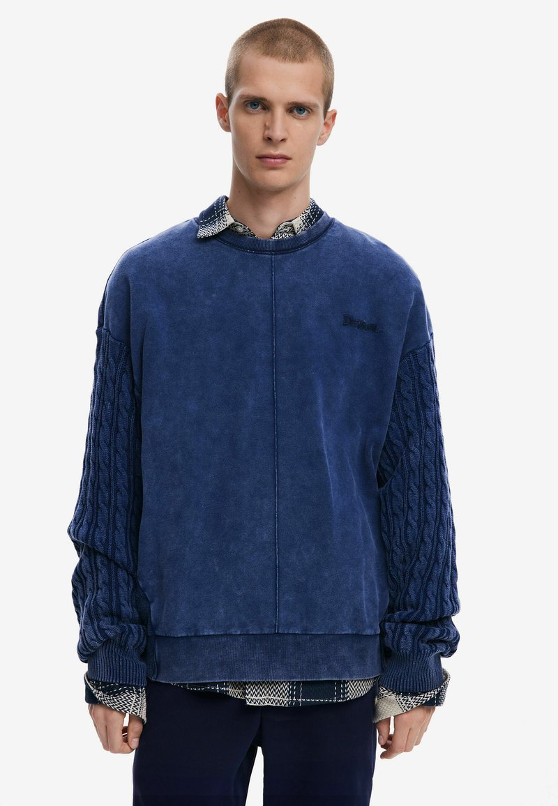 Blue oversized sweatshirt with textured cable knit sleeves, casual fit, and a ribbed cuff. Layered over a checkered shirt.