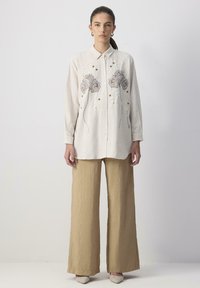 Light beige button-up shirt with floral embroidery in blue and gold, paired with wide beige trousers and cream-colored shoes.