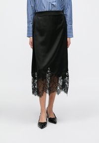 Zwarte satijnen rok met een gegolfde kanten zoom, voorzien van ingewikkelde bloemmotieven. Gecombineerd met zwarte puntige hakken en een blauw-gestreept topje.
