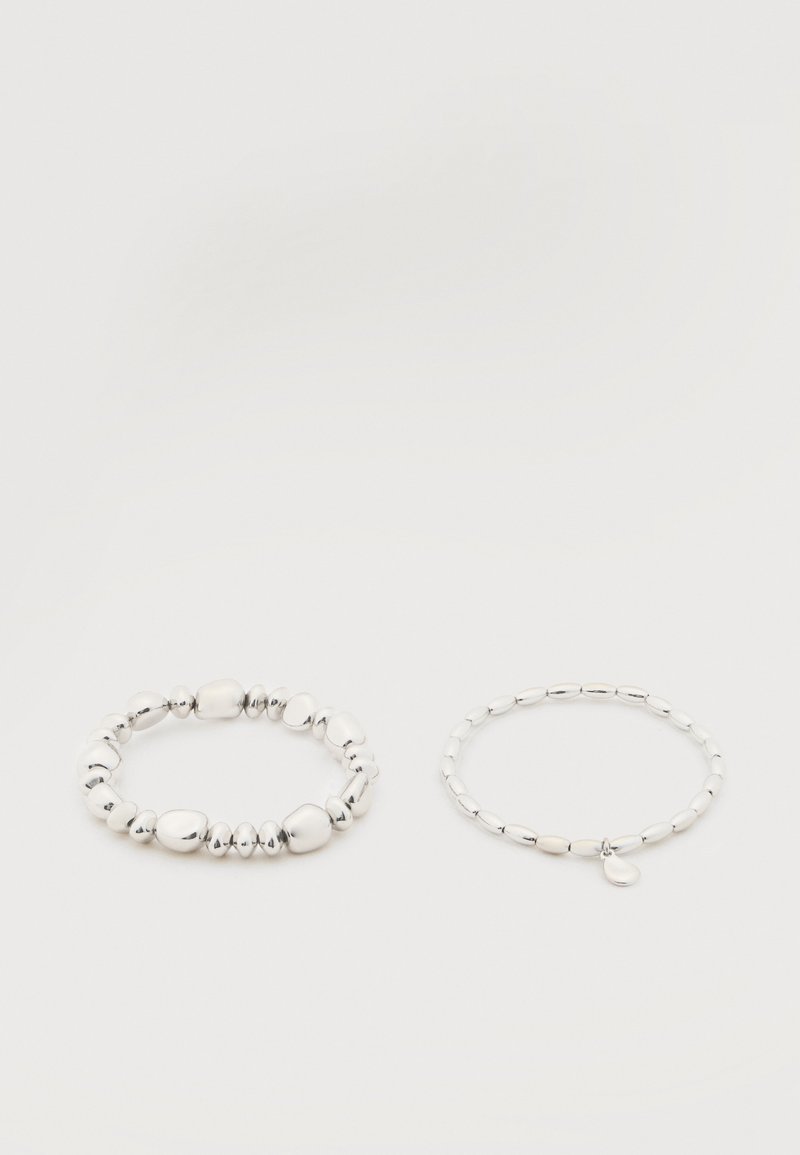 Deux bracelets de perles argentées sur fond blanc ; l'un avec des perles irrégulières et l'autre avec des perles uniformes et un petit charme en forme de goutte.