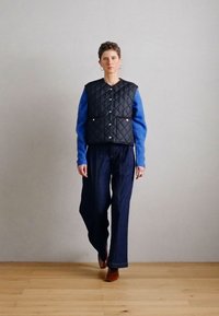 Pihegett fekete mellény, kék hosszú ujjú felső, sötét farmernadrág, barna bokacsizma. Az öltözék kontrasztos textúrákat és színeket mutat.