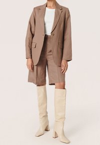 Donna che indossa un completo giacca e pantaloni corti marrone con un top bianco a costine e stivali alti color crema con tacco che si trova su uno sfondo neutro.