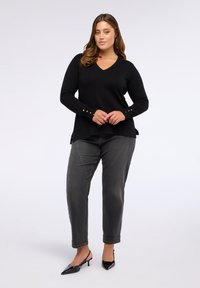 Suéter negro de punto con escote en V, mangas largas y detalles de botones dorados en los puños, combinado con jeans grises y tacones negros de punta.