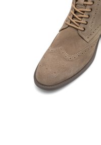 Stivaletto brogue beige in suede con punta rotonda, dettagli perforati, lacci beige e suola in gomma. Inquadratura ravvicinata che si concentra sulla costruzione superiore.
