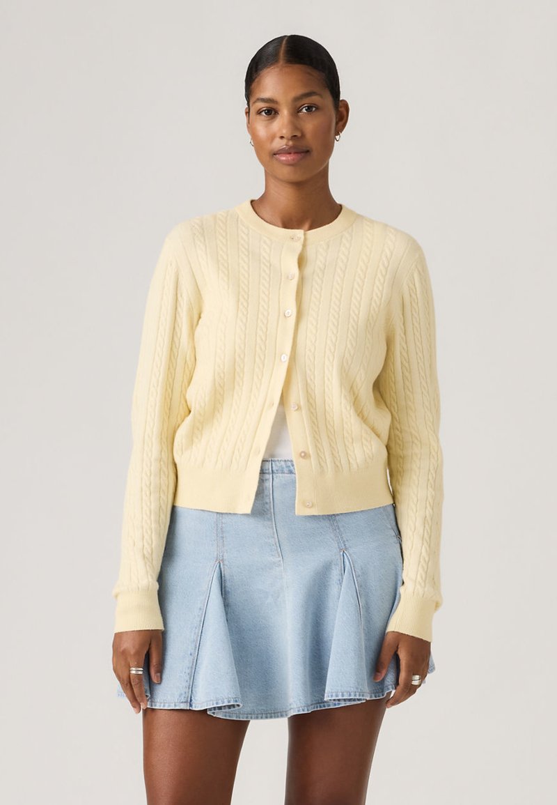 Γυναίκα που φοράει ανοιχτό κίτρινο πλεκτό ζακέτα με κουμπιά και ανοιχτό μπλε πλισέ τζιν φούστα, στέκεται μπροστά σε απλό φόντο.