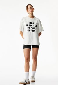 Valkoinen ylisize-paita, jossa on rohkea musta teksti "EN INSPIROITU TÄNÄÄN, NÄIN PÄÄLLÄ." Yhdistetty mustiin shortseihin ja lenkkareihin.