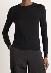 Personne portant une chemise noire ajustée à manches longues et un pantalon cargo foncé, avec une main dans la poche et un fond neutre.