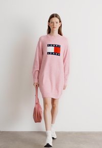Roza oversize pulover obleka z rebrasto teksturo, z logotipom v mornarsko modri, rdeči in beli barvi. Model nosi usklajen roza torbico čez ramo.