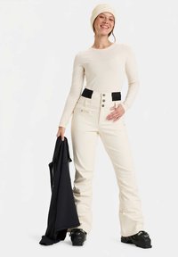 Mlada žena u svijetlosmeđem skijaškom odijelu i kapici drži crnu jaknu, nosi crne skijaške čizme i smiješi se na bijeloj pozadini.