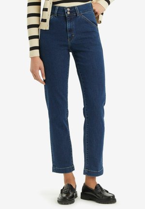 Kobieta w ciemnoniebieskich spodniach jeans o wysokim stanie z podwójnymi guzikami, podwiniętymi nogawkami, czarnych mokasynach oraz w paskowanym kremowo-czarnym bluzce z długim rękawem.