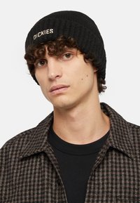 Černá pletená čepice s ribbenou texturou a vyšívaným logem "DICKIES". Nositá s kostkovanou flanelovou košilí přes černé tričko s kulatým výstřihem.