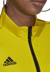 Žuta jakna na zip s crnim patentnim zatvaračem i Adidas logotipom na donjem lijevom dijelu. Tkanina izgleda glatko s blagom teksturom.