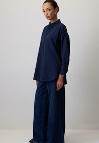 Camicia blu navy con bottoni, maniche lunghe e orlo curvo, abbinata a jeans blu a gamba larga. Design semplice, texture liscia.