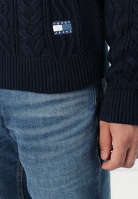 Marineblauwe gebreide trui met gestructureerde kabelpatronen, geribde manchetten en zoom, beschikt over een klein logopatch. Gekocht met lichtblauwe jeans.