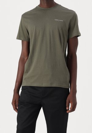 Mand iført olivengrøn kortærmet Tommy Jeans t-shirt og sorte bukser, stående mod en ensartet lys baggrund.