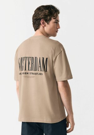 T-shirt en coton beige avec une coupe décontractée, présentant une grande impression noire sur le dos disant "AMSTERDAM" avec un texte plus petit en dessous.
