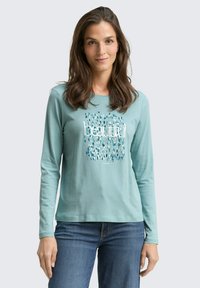 Woman with long brown hair wearing teal long-sleeve shirt with "beautiful" text and abstract design, paired with blue jeans, standing against plain background.