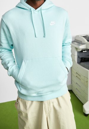 Persoon in een lichtblauwe Nike-hoodie en beige broek staat bij een kantoorprinter op een groen tapijt.