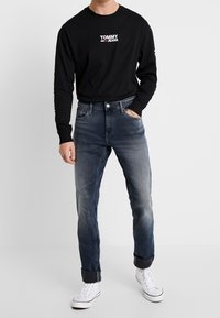 Svart långärmad skjorta med "TOMMY JEANS" logotyp, kombinerad med blekta blå denimjeans och vita sneakers. Upprullade muddar på jeansen.