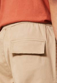 Pantalones beige con un acabado texturizado, con una cintura elástica y un bolsillo trasero sin herrajes visibles. El color de la camisa es óxido.