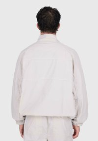 Giacca leggera di colore chiaro, con colletto alto, polsini elasticizzati e due cuciture orizzontali sulla schiena. Texture liscia e opaca.