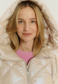 Giacca a vento imbottita di un beige chiaro con una texture lucida e trapuntata. Presenta una chiusura con zip e un ampio colletto con cappuccio. Indossata sopra un maglione rosa.