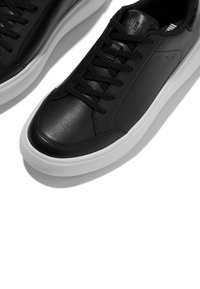 Zapatillas deportivas bajas de cuero negro con cordones negros y suelas gruesas de goma blanca sobre un fondo blanco.