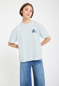 Jeune femme debout, les mains derrière le dos, portant un t-shirt ample bleu clair avec un petit graphisme coloré et un jeans large bleu.