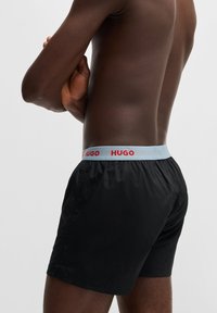 Sorte bokserer med elastisk talje, der har en lys blå kant og rød "HUGO" logo. Glat tekstur og tætsiddende design.