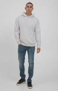 Helles graues Hoodie mit einer Kängurutasche und gesticktem Text, kombiniert mit blauen Jeans und schwarzen Sneakers, im schlanken Schnitt und mit subtilen Mängeln.